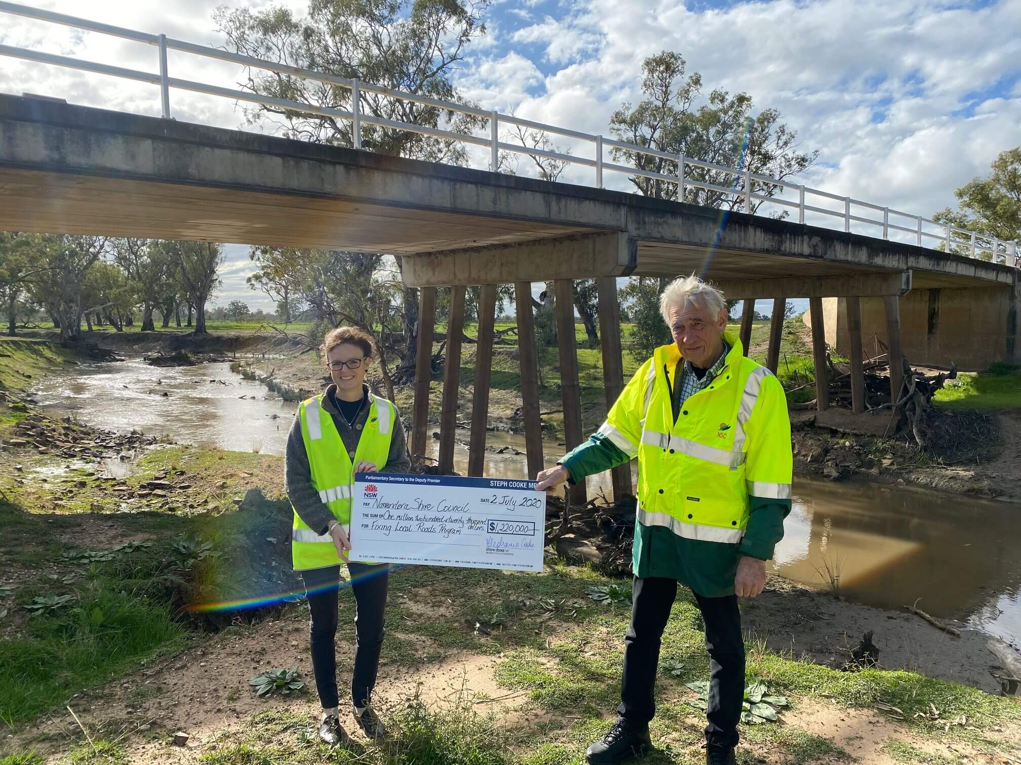 teph Cooke and Narrandera Mayor Neville Kschenka at the Brewarrina Bridge. They hold a large cheque between them and wear hi-vis vests. The underside of the bridge can be seen.