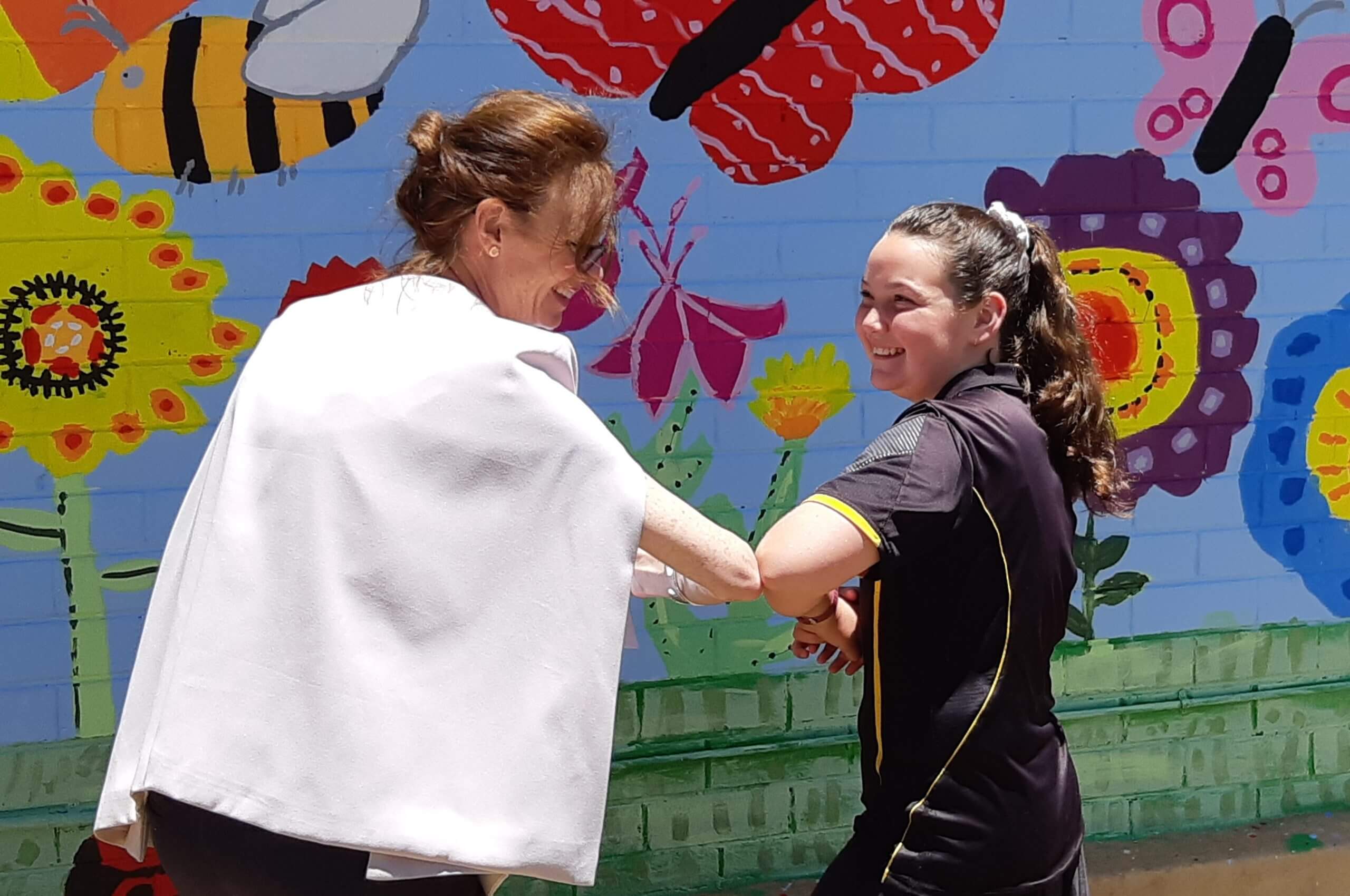 Steph Cooke and a female student bump elbows in front of a brightly painted wall with large flowers, butterflies and bees.