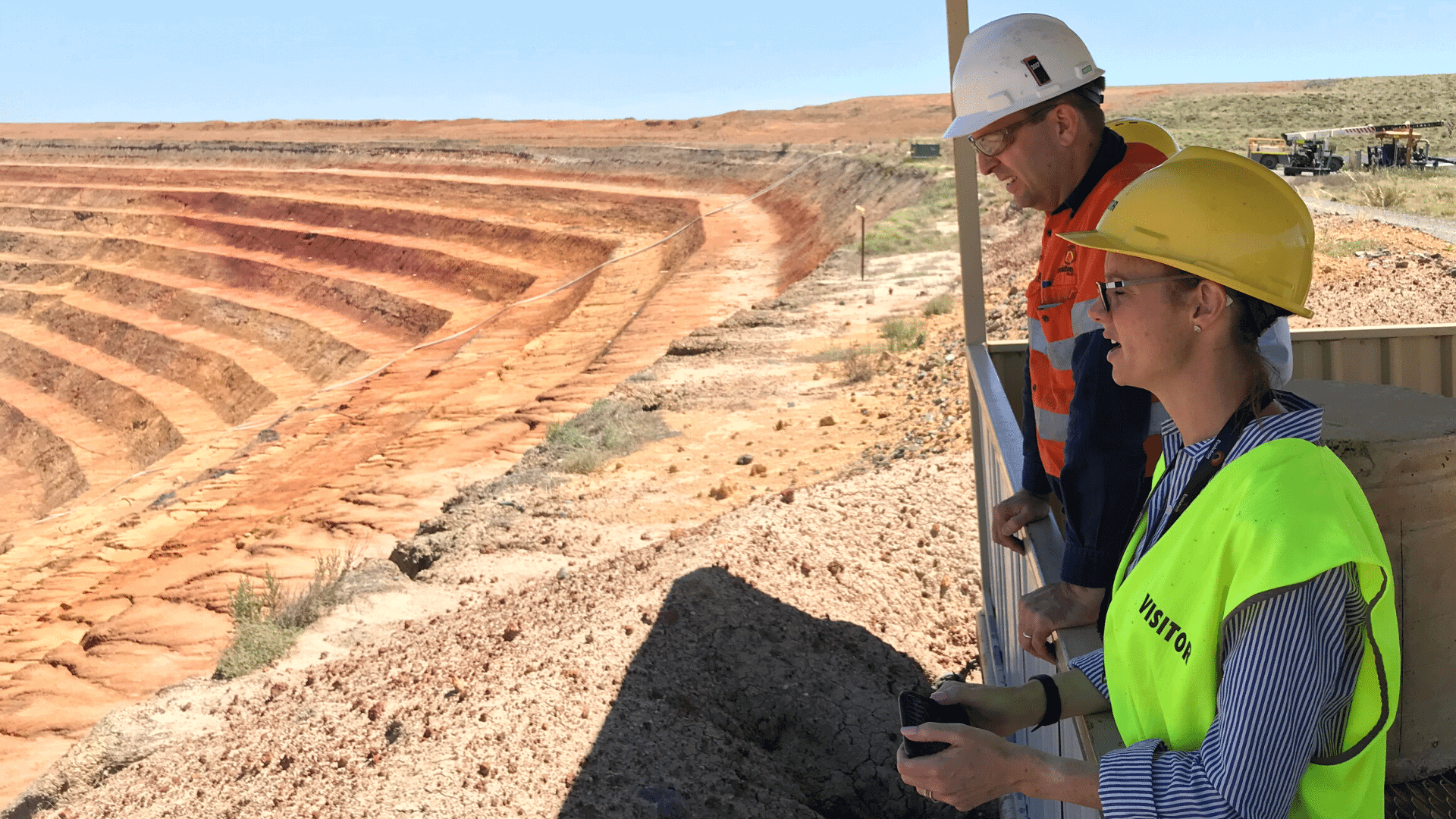Member for Cootamundra Steph Cooke MP prior to social distancing inspects Lake Cowal Gold Mine in the Bland Shire with visitor high viz vest on and hard hat. The golden stacked quarry is visible in the background as a mining representative also looks on beside Steph.