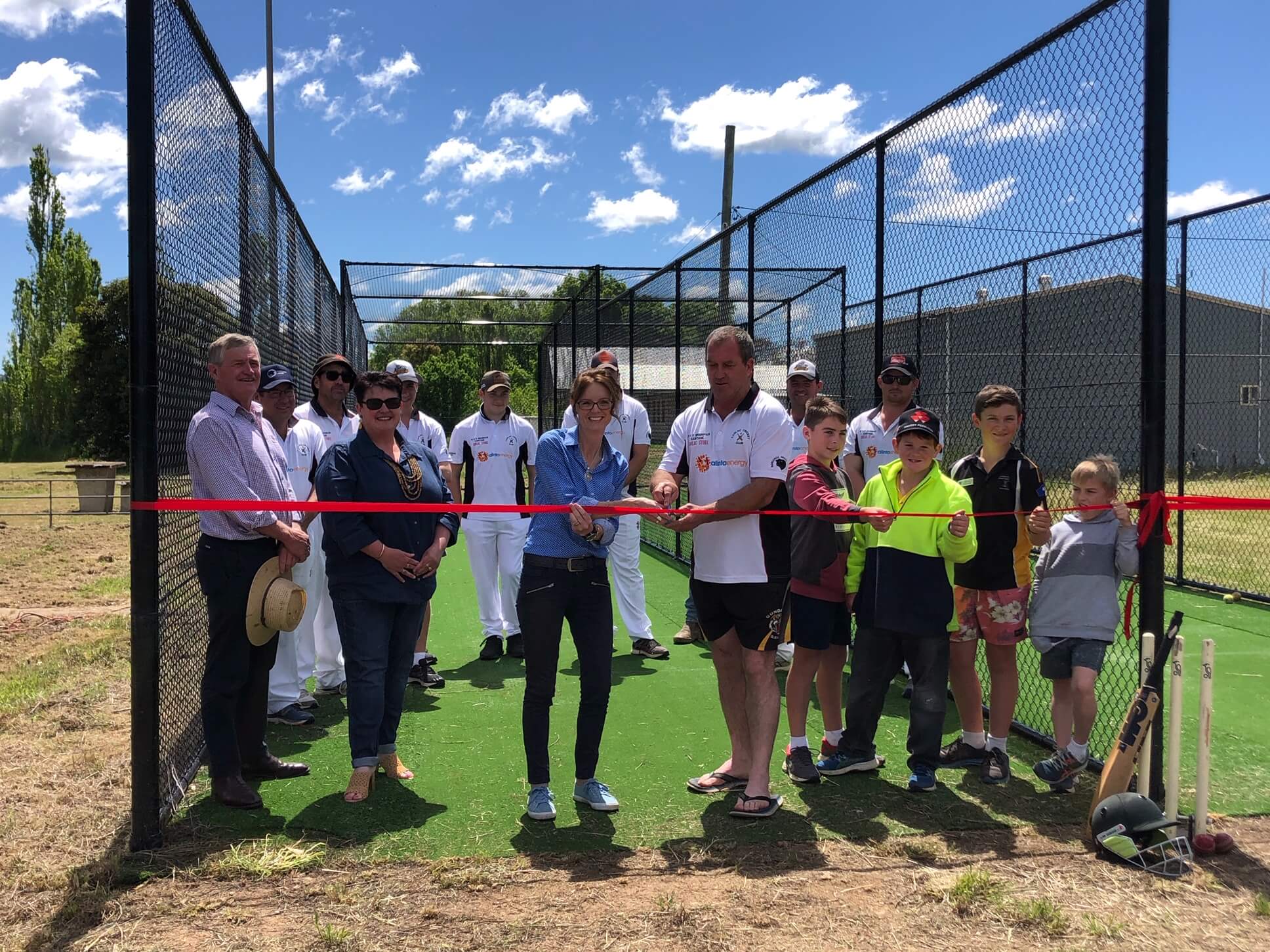 Steph Cooke stands in the new nets and cuts a red ribbon. Holding the ribbon is Andrew Scott who wears a white cricket polo shirt. Surrounding them are adults and children from the Coolac Cricket Club.