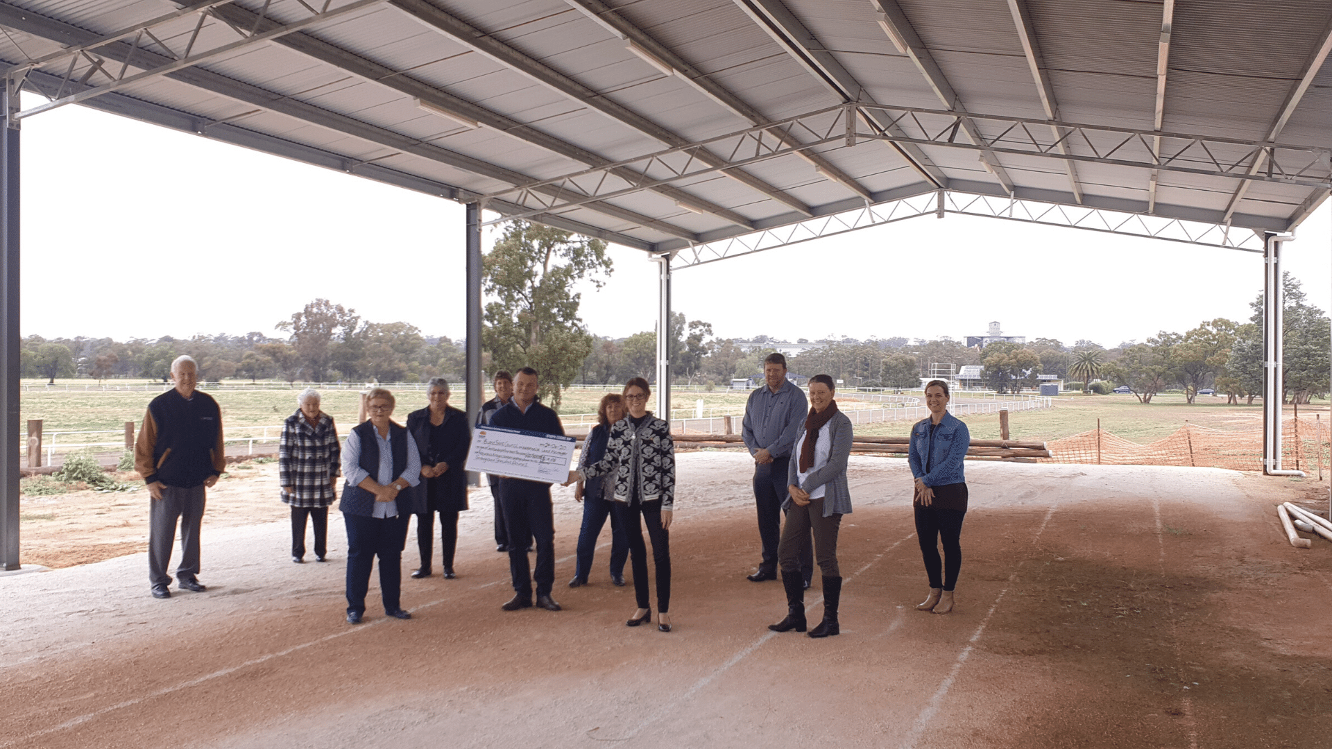 Steph Cooke MP stands in middle front row wearing a black and white coat with black pants holding a cheque with Craig Sutton, and Representatives of Weethalle community and Bland Shire Council under a large steel structure overlooking rural landscape