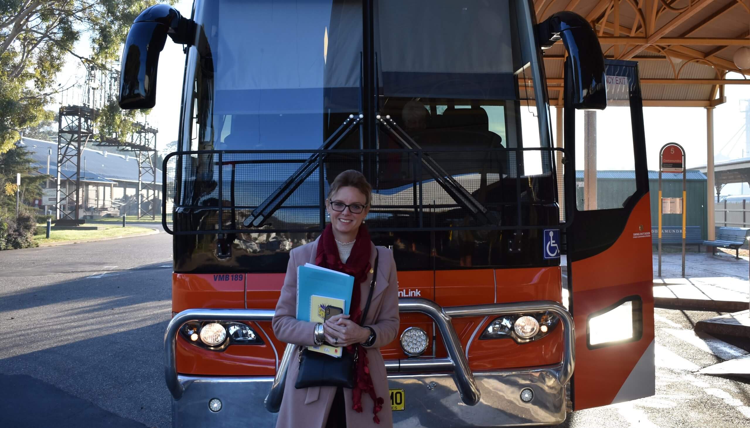 Steph Cooke MP stands in front of a red bus wearing caramel coat and red scarf whilst holding documents and folders. The bus is parked outside a bus shelter with some steel infrastructure in background