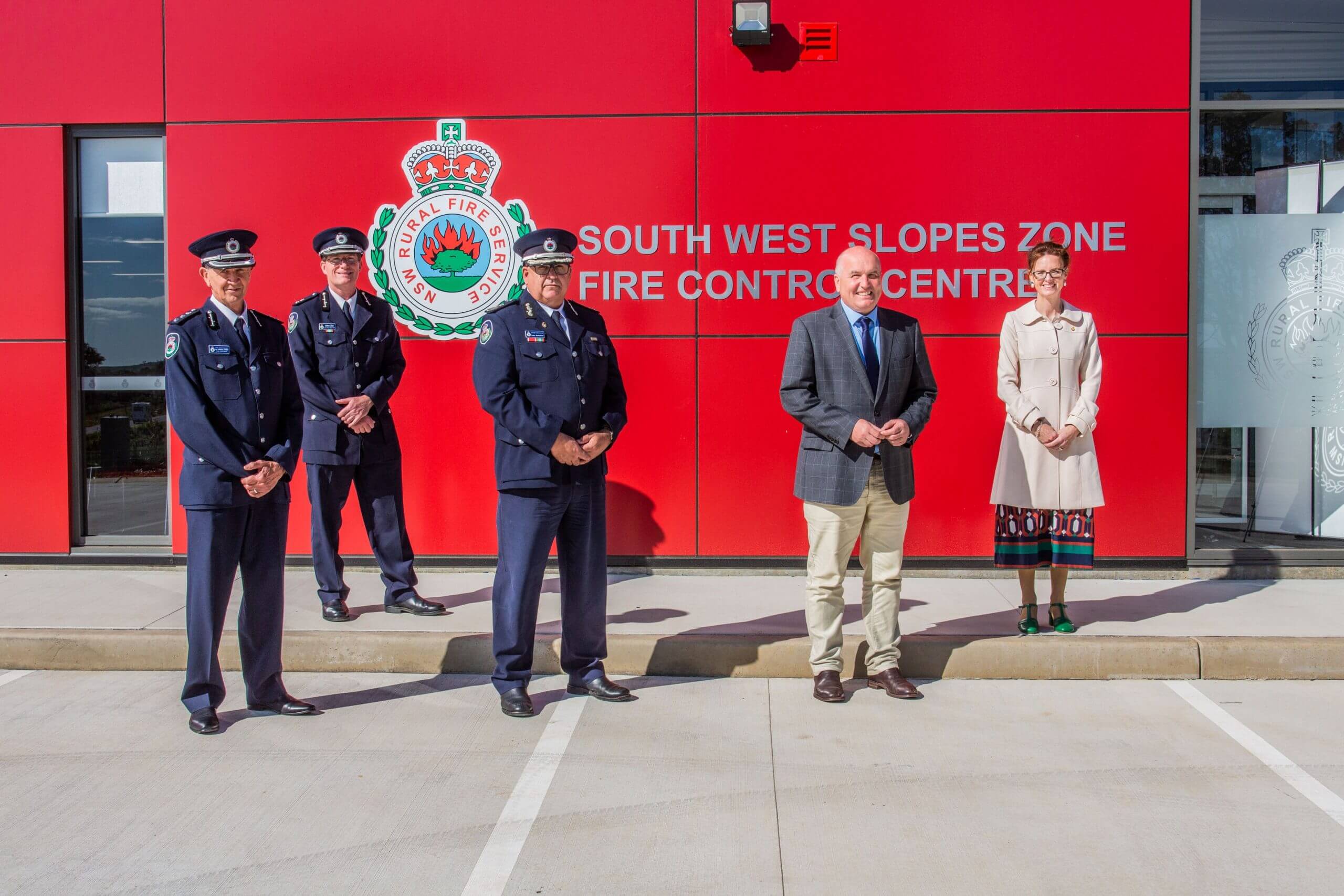 RFS Representatives, Minister Elliott and Steph Cooke stand in front of the bright red front of the fire control centre.