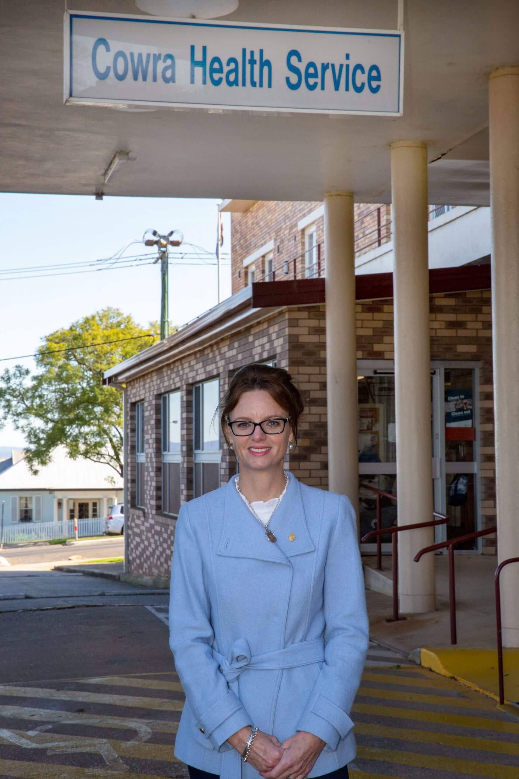 Steph Cooke stands under the sign for the Cowra Health Service. She wears a pale blue jacket and smiles at the camera. The hospital entrance can be seen in the background.