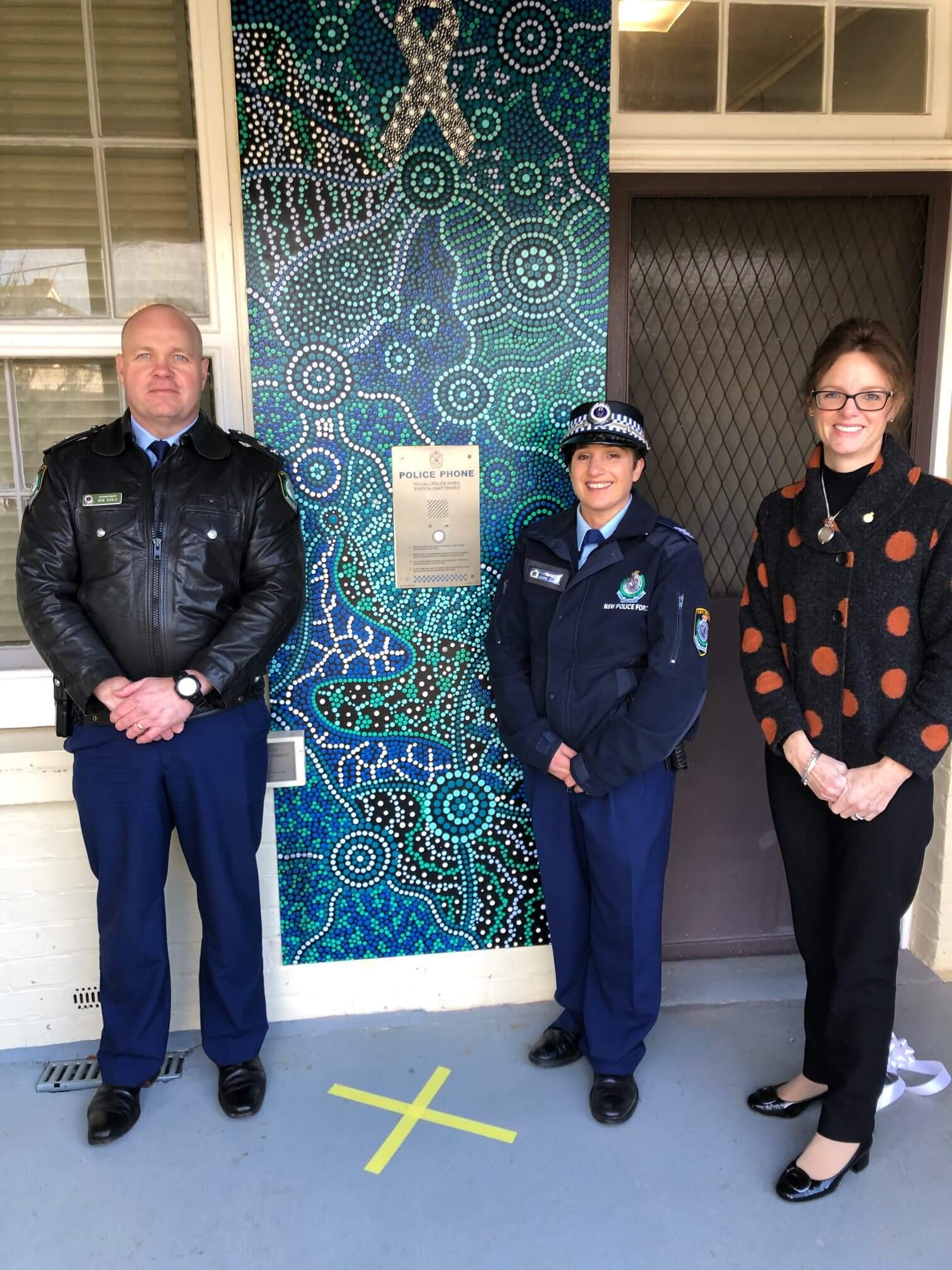Superintendent Bob Noble, Sergeant Joanne Gallant and Steph Cooke MP stand in front of the blue, black and white indigenous artwork which features a prominent white ribbon