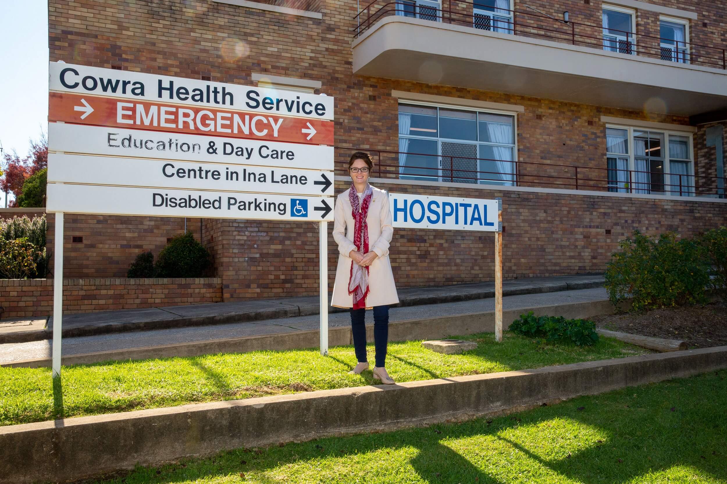 Steph Cooke stands next to a sign that reads 'Cowra Health Service' and 'emergency'. The hospital building is behind her.